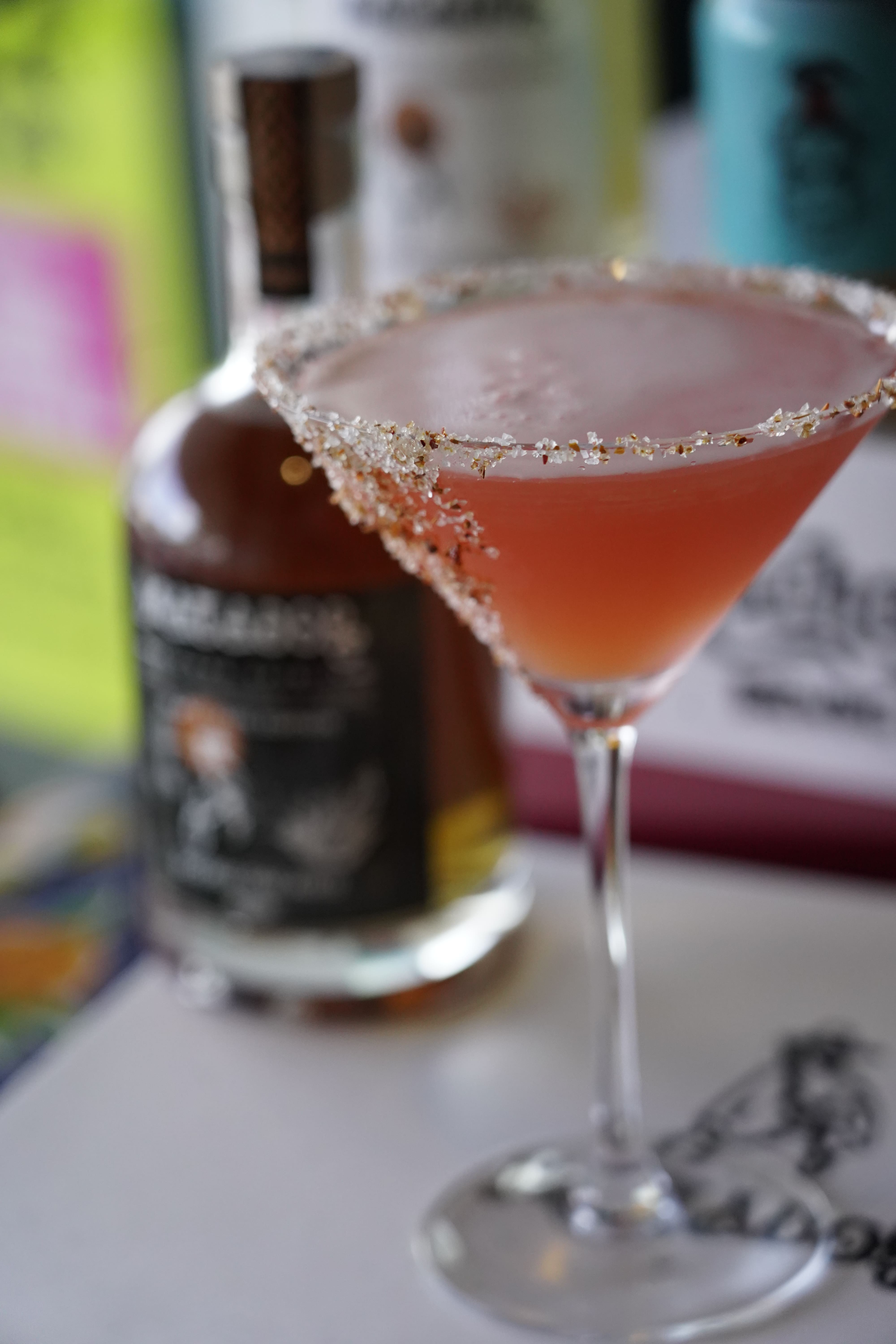 Pink cocktail in a martini glass with a sugared rim, surrounded by bottles on a table. Margys