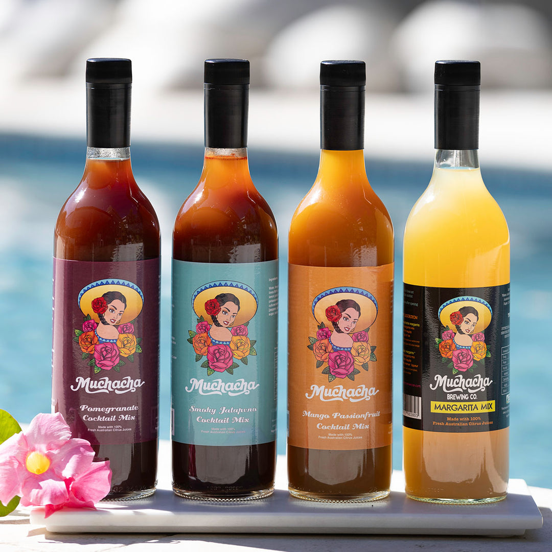 Four bottles of Muchacha cocktail mix with colorful labels on a white surface by a pool.