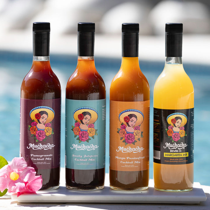 Four bottles of Muchacha cocktail mix with colorful labels on a white surface by a pool.