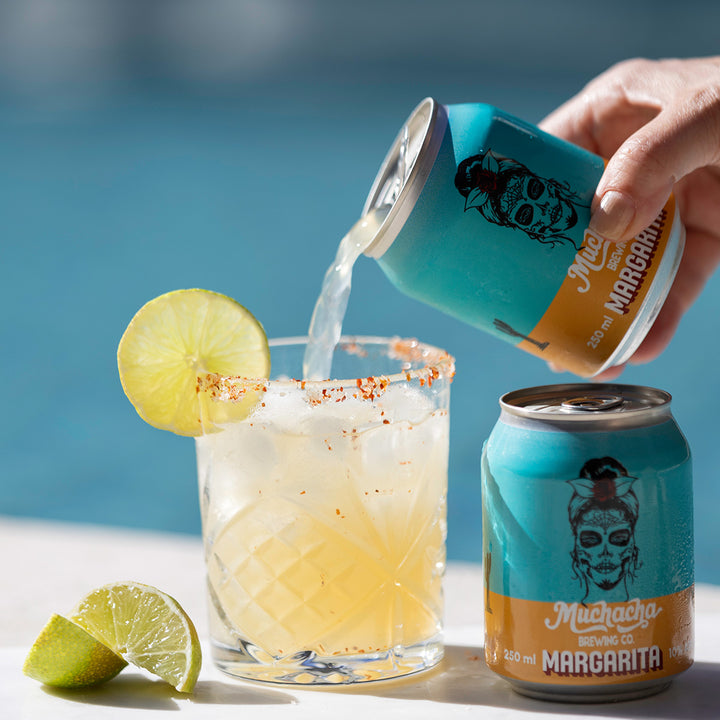 Person pouring a Margarita from a can into a glass with lime slices, with another can of Margarita in the background.